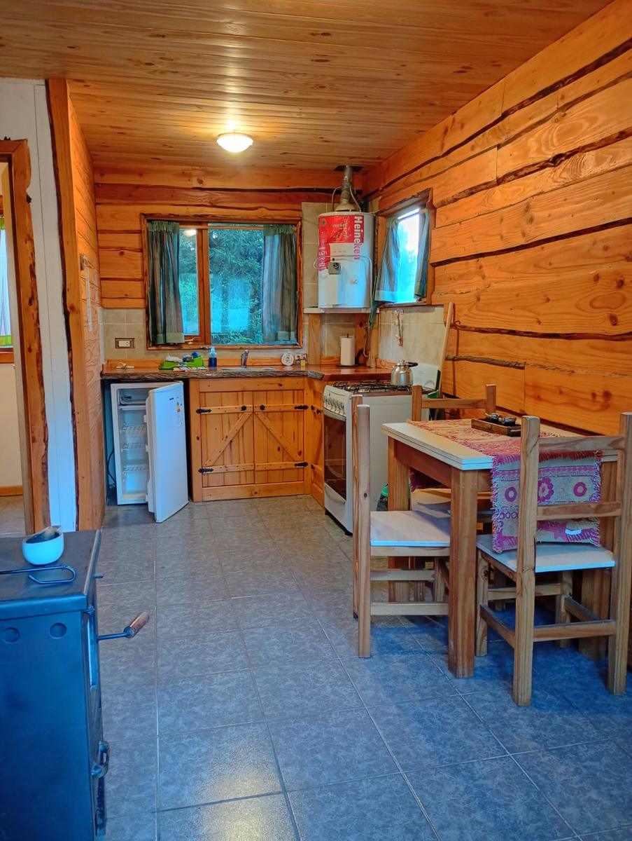 Cocina y comedor de la cabaña, con cocina a gas, heladera, mesa de madera y la estufa a leña